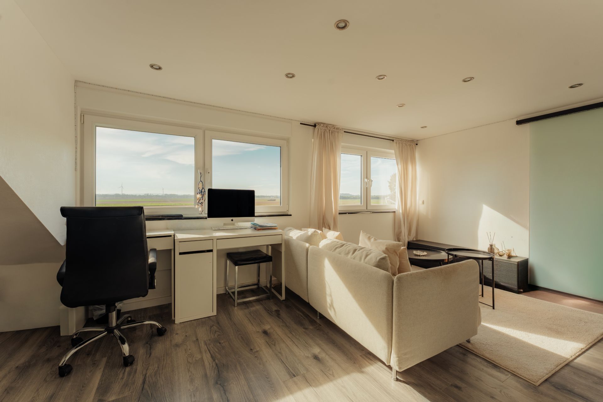 Helles Wohnzimmer mit Schreibtisch, Sofa und großem Fenster, Parkettboden.