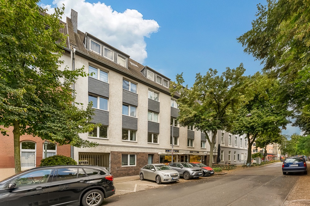 Mehrstöckiges Wohngebäude mit grauer Fassade, geparkte Autos und Bäume an der Straße.