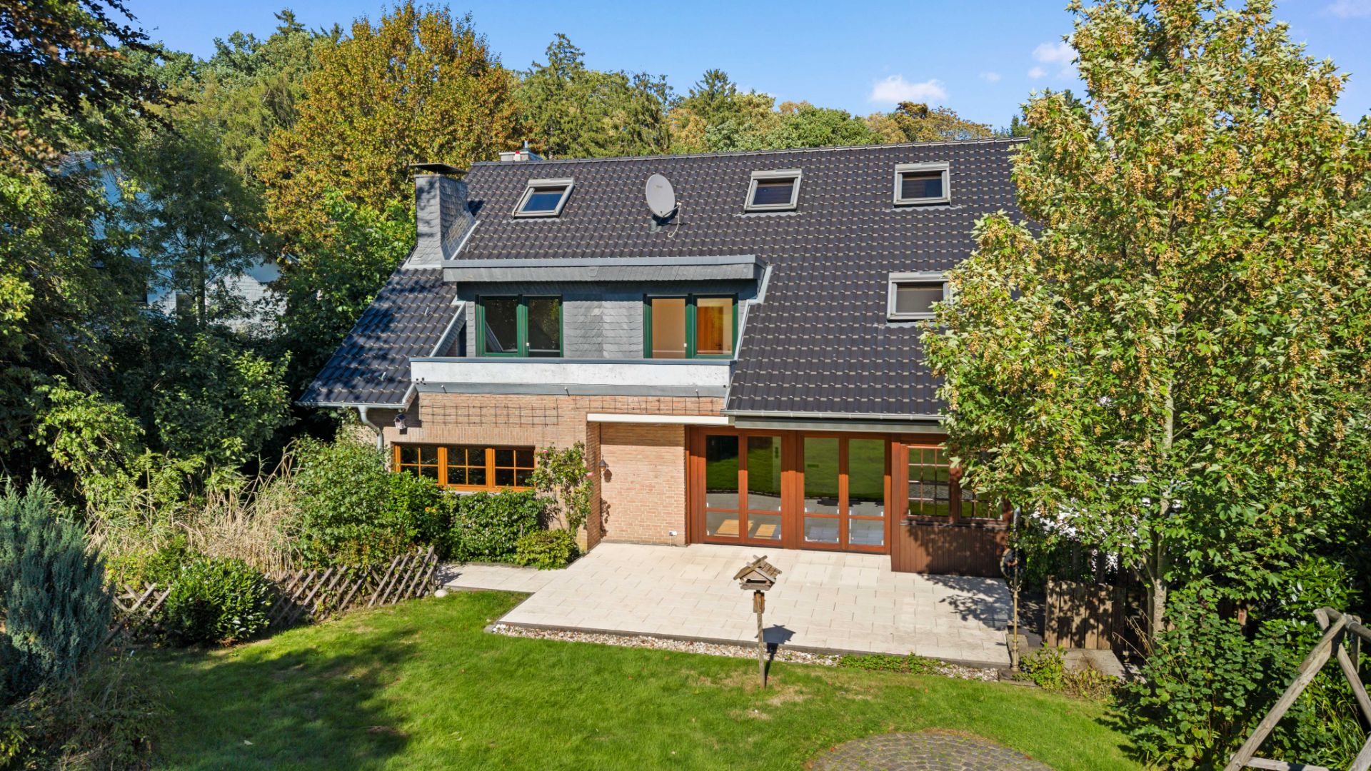 Zweistöckiges Wohnhaus mit dunklem Dach, Terrasse und großem Garten, von Bäumen umgeben.