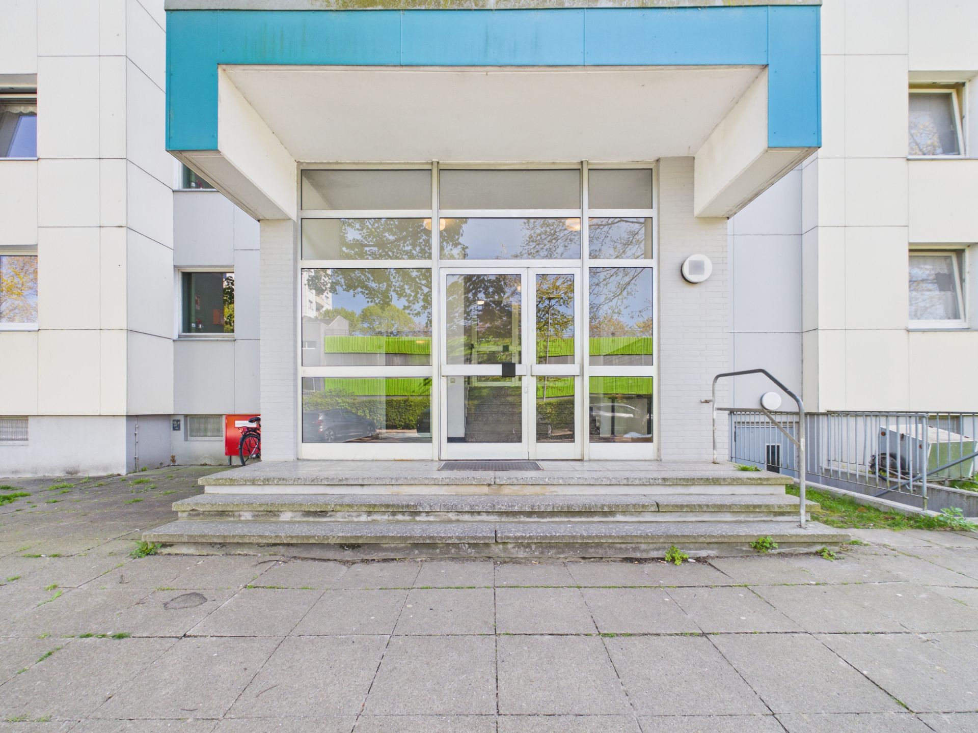 Eingang eines Wohngebäudes mit Glastür, Stufen und blauem Vordach.