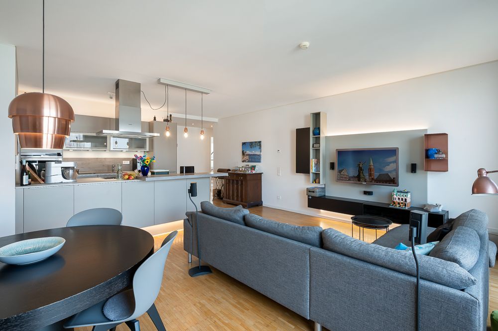 Modernes Wohnzimmer mit grauem Sofa, Esstisch und offener Küche mit Hängelampen.