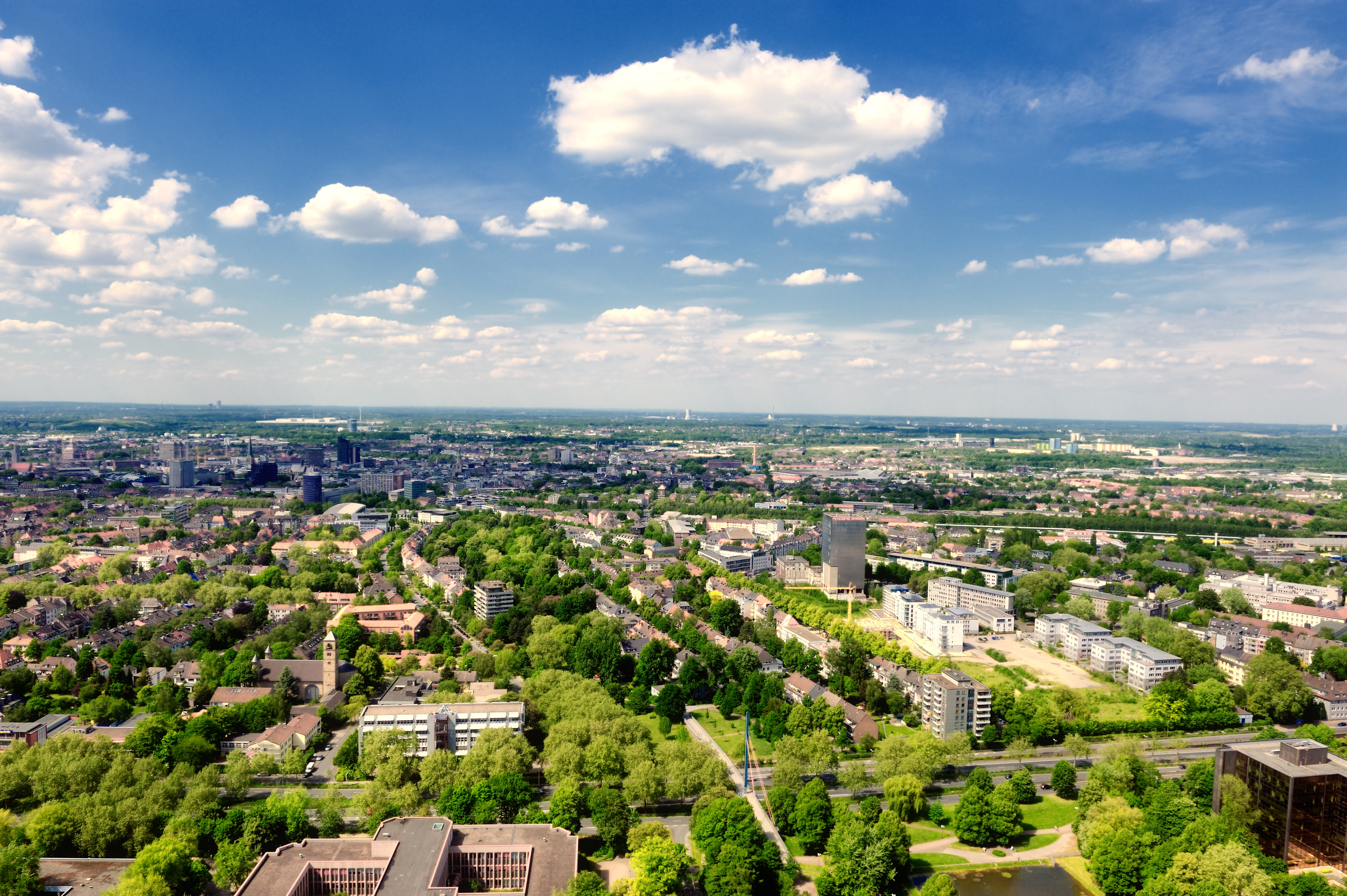 Blick von oben auf eine Stadt mit vielen Bäumen, Wohngebäuden und blauem Himmel mit Wolken.