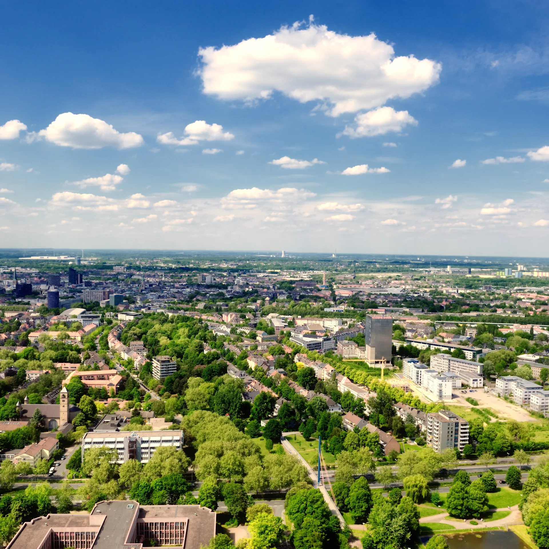 Blick von oben auf eine Stadt mit vielen Bäumen, Wohngebäuden und blauem Himmel mit Wolken.