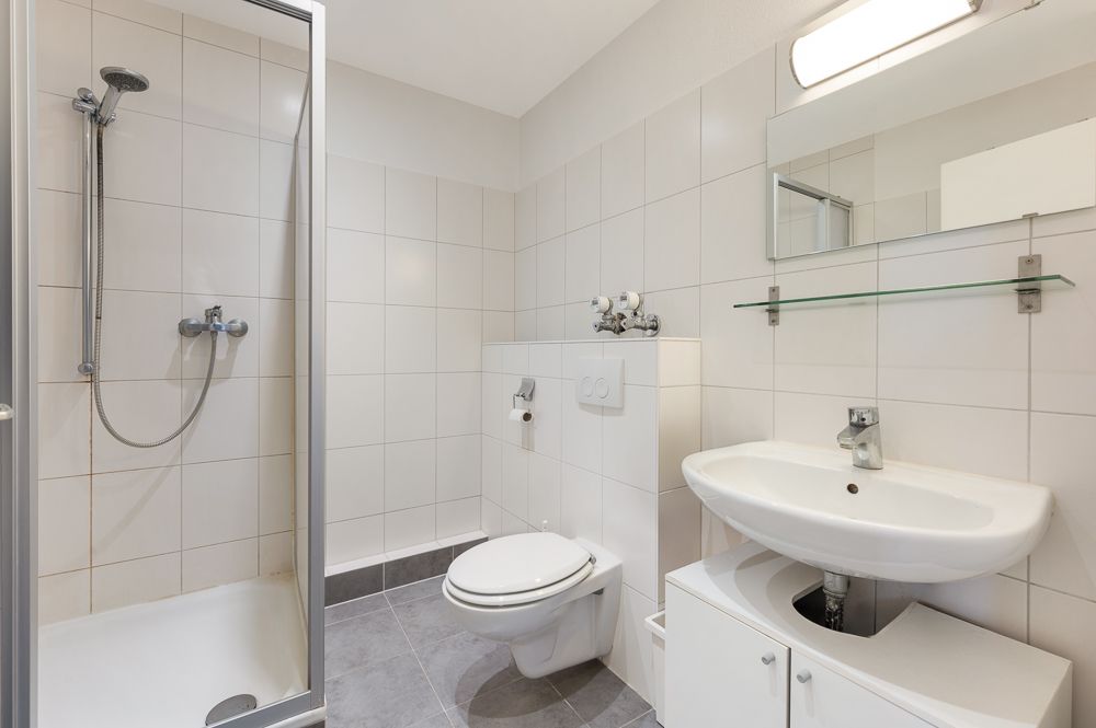 Helles Badezimmer mit Dusche, Toilette, Waschbecken, Spiegel und Glasablage.