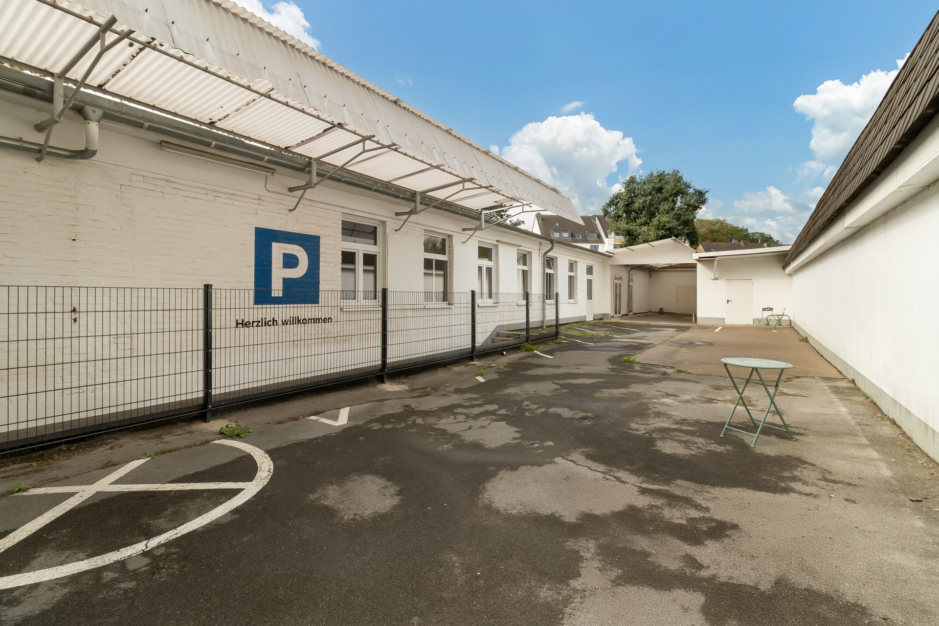 Gepflasterter Hof mit Parkplatzmarkierung, weißem Gebäude und kleinem Klapptisch neben dem Zaun.