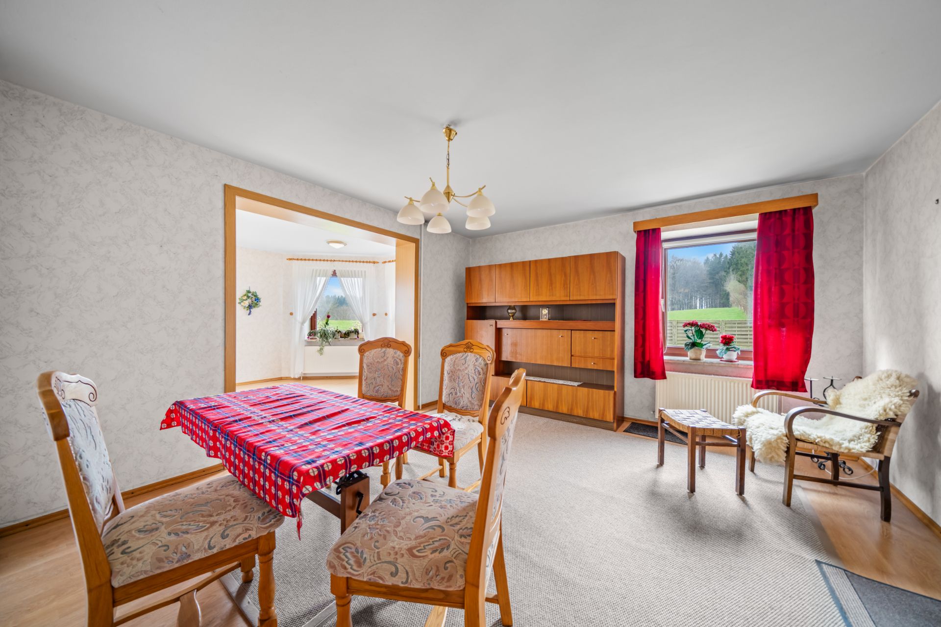 Helles Wohnzimmer mit Esstisch, vier Stühlen, Sideboard, Sessel und roten Vorhängen am Fenster.
