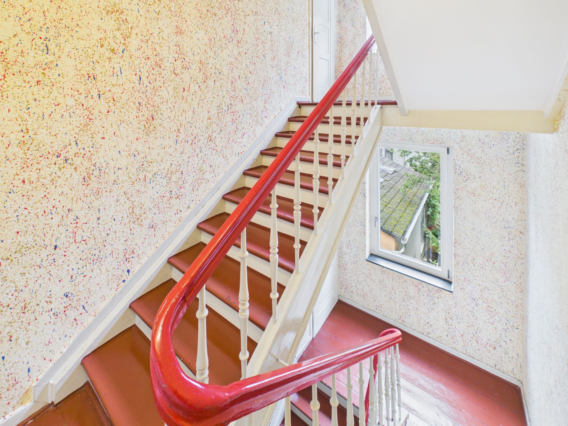 Treppenhaus mit rotem Handlauf, bunter Wand und Fenster, durch das man ins Freie sieht.