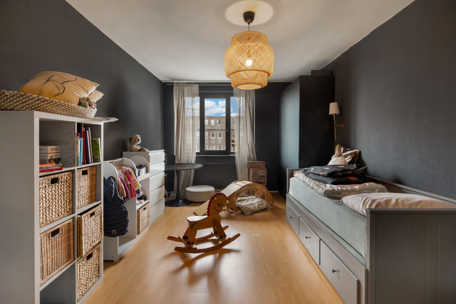 Kinderzimmer mit Spielzeugpferd, Regal mit Körben, Bett und großem Fenster.