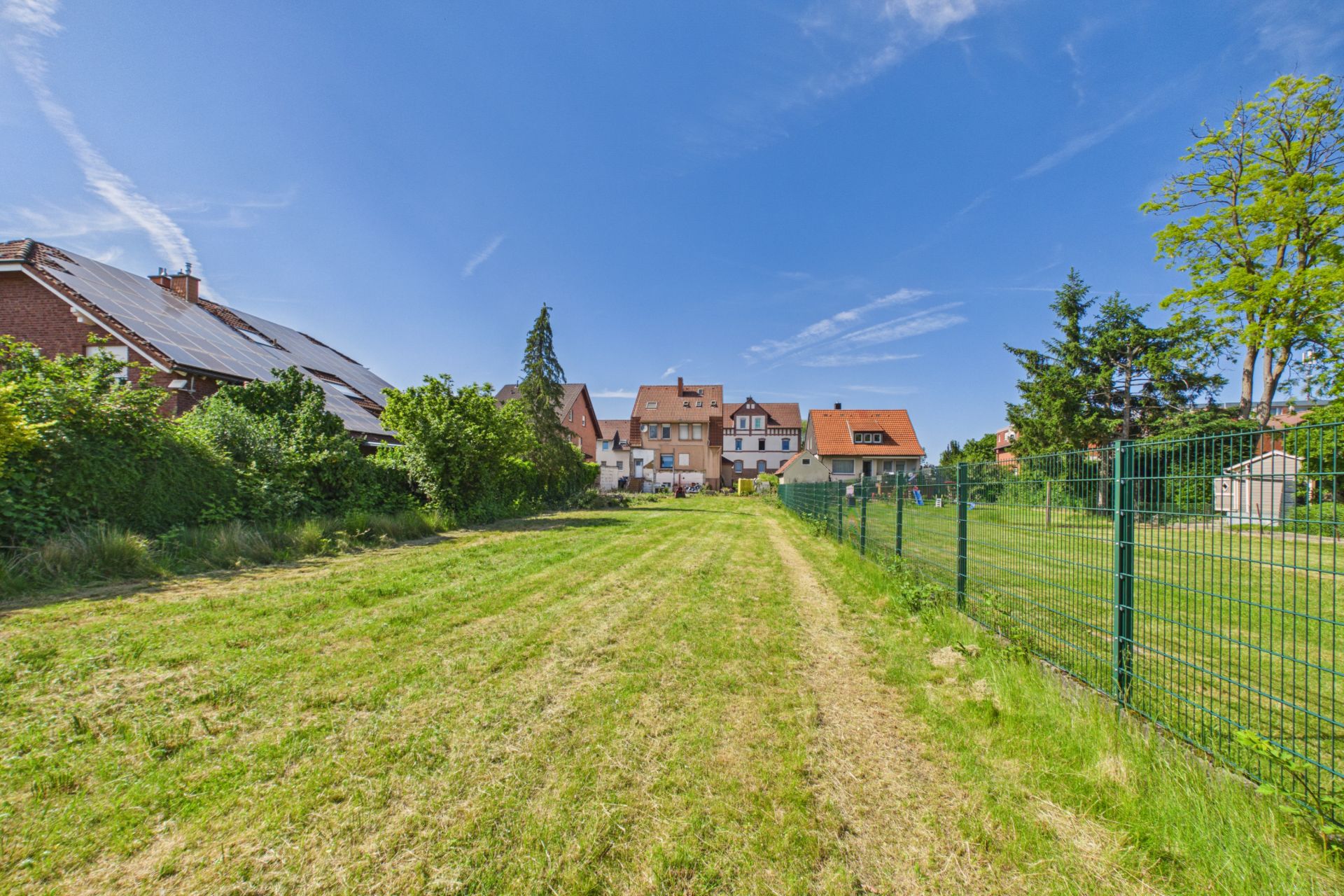 Großer, gemähter Garten mit Blick auf Häuser im Hintergrund, eingezäunt und von Bäumen umgeben.