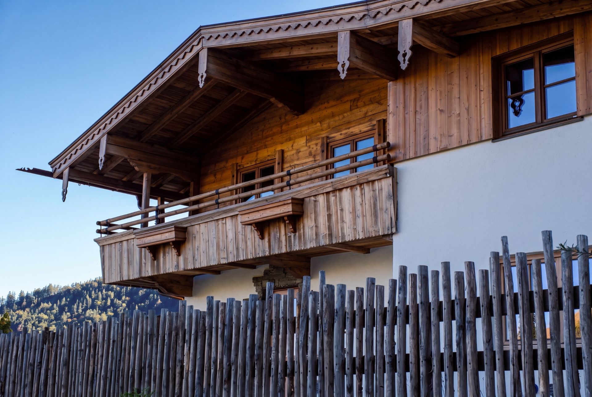 Holzhaus mit Balkon und Holzzaun im Vordergrund, im Hintergrund Bäume und blauer Himmel.