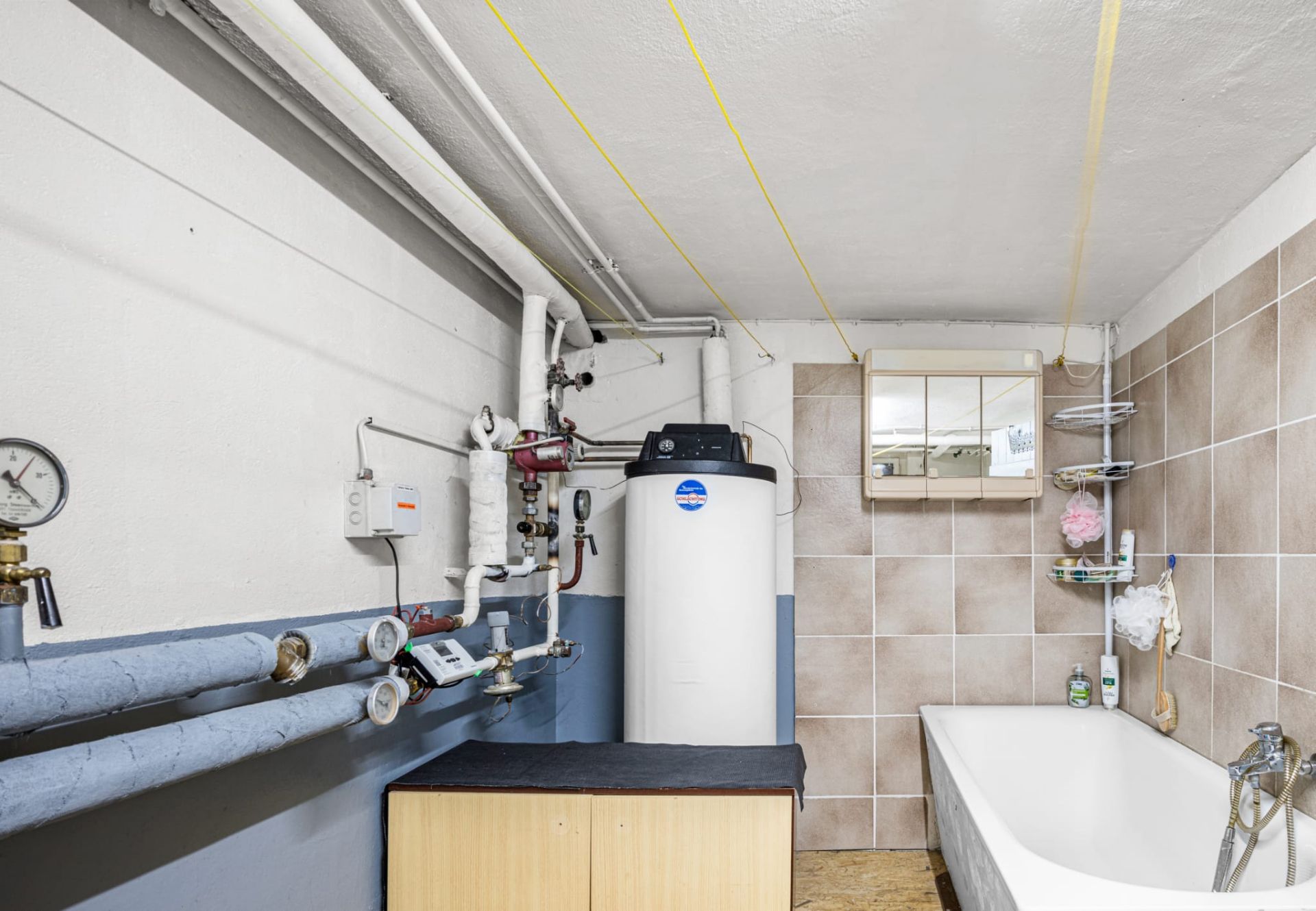 Heizungsraum mit Rohren, Warmwasserspeicher, Badewanne und gefliester Wand.