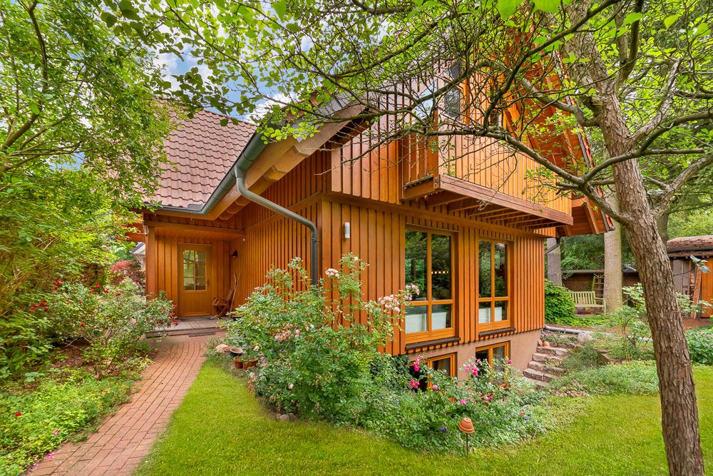 Holzhaus mit großen Fenstern, Garten und Bäumen, Weg führt zum Eingang.