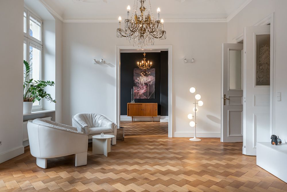 Helles Wohnzimmer mit zwei weißen Sesseln, Parkettboden, Kronleuchter und Stehlampe.