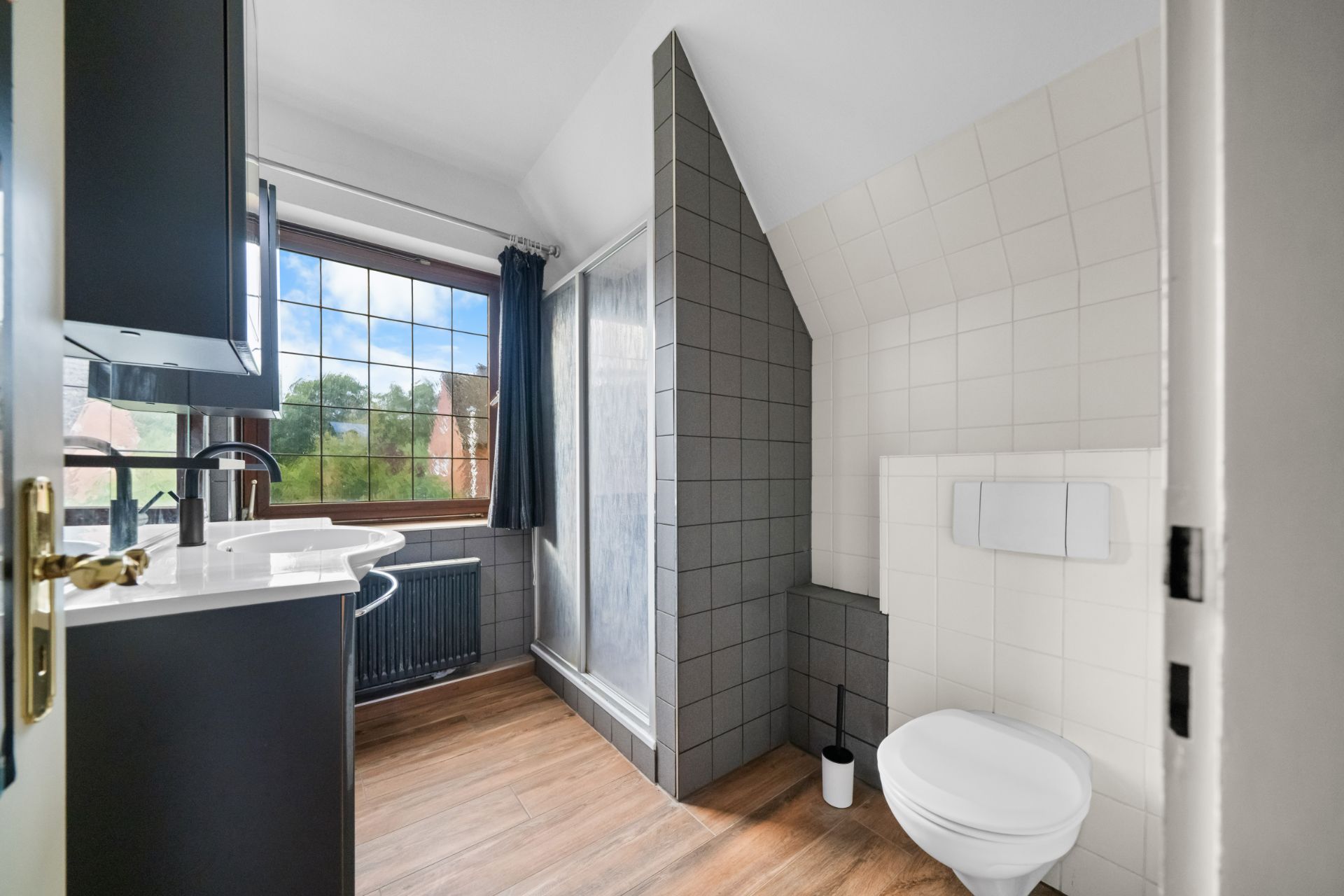 Helles Badezimmer mit Dusche, Waschbecken und Wandtoilette, Holzboden und Fenster.