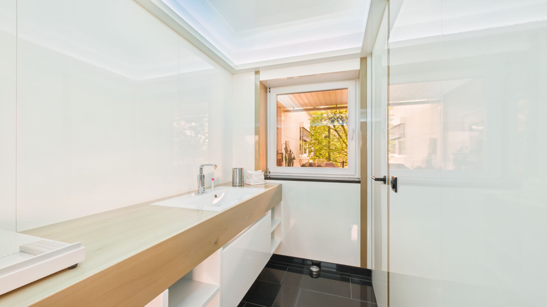 Modernes Badezimmer mit Waschbecken, Spiegel und Fenster, weiße Wände und schwarze Fliesen.