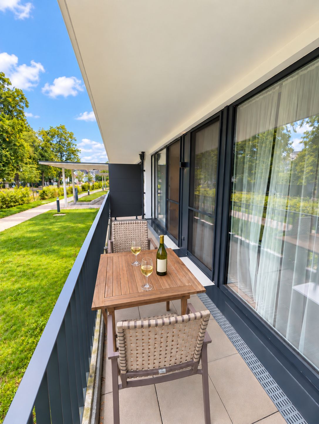 Balkon mit Holztisch, zwei Stühlen und einer Flasche Wein sowie zwei Gläsern, Blick auf einen Garten.