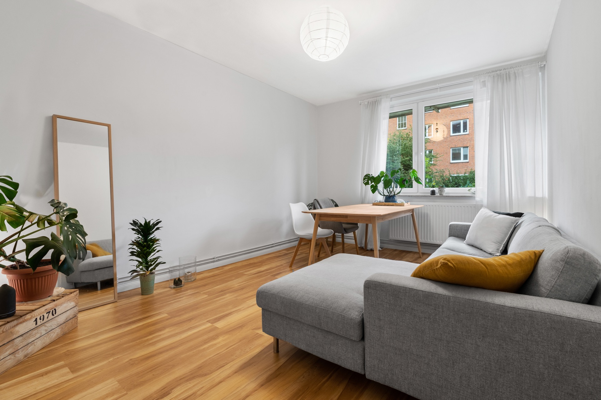 Helles Wohnzimmer mit grauem Sofa, Holzboden, Tisch, zwei Stühlen, Pflanzen und großem Fenster.