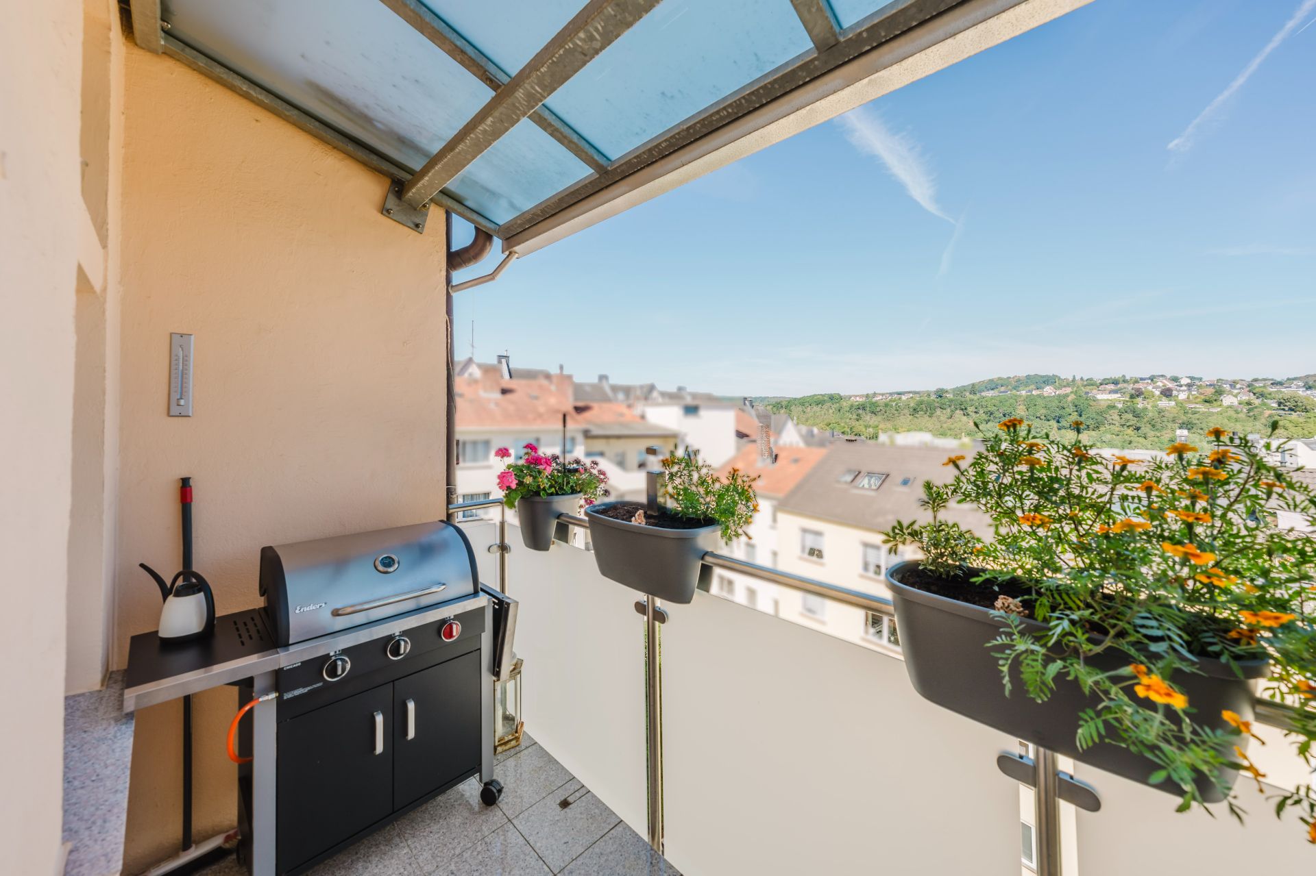 Balkon mit Grill, Pflanzen in Blumenkästen und Blick auf Häuser und grüne Hügel.