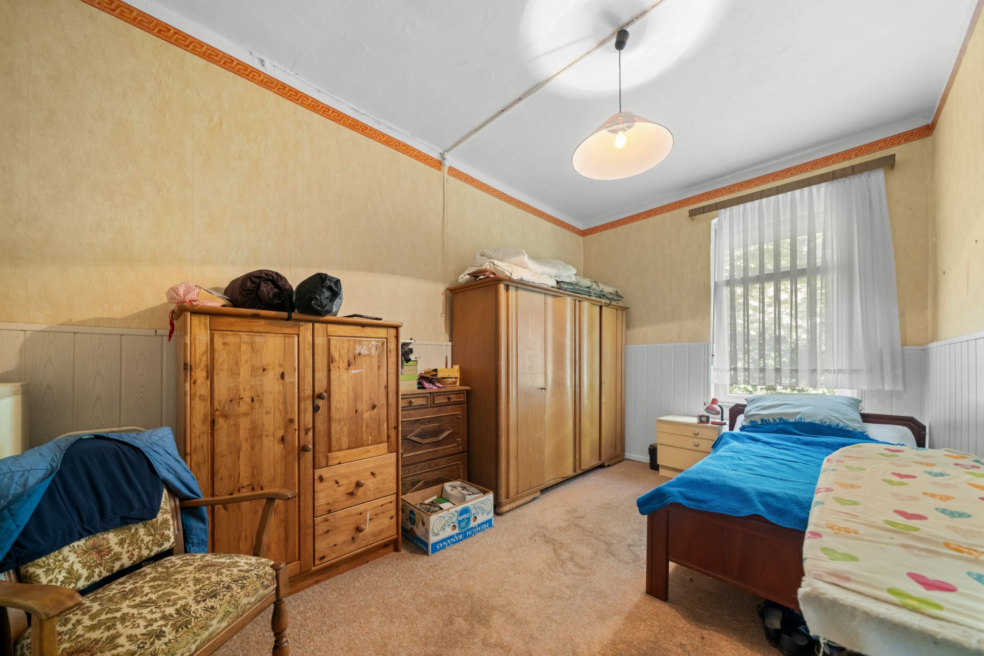 Schlafzimmer mit zwei Betten, Holzschränken, Kommode, Stuhl und großem Fenster mit Gardinen.