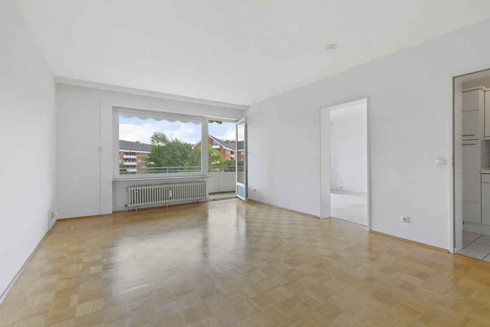 Helles Wohnzimmer mit Parkettboden, großem Fenster, Zugang zum Balkon und angrenzender Küche.