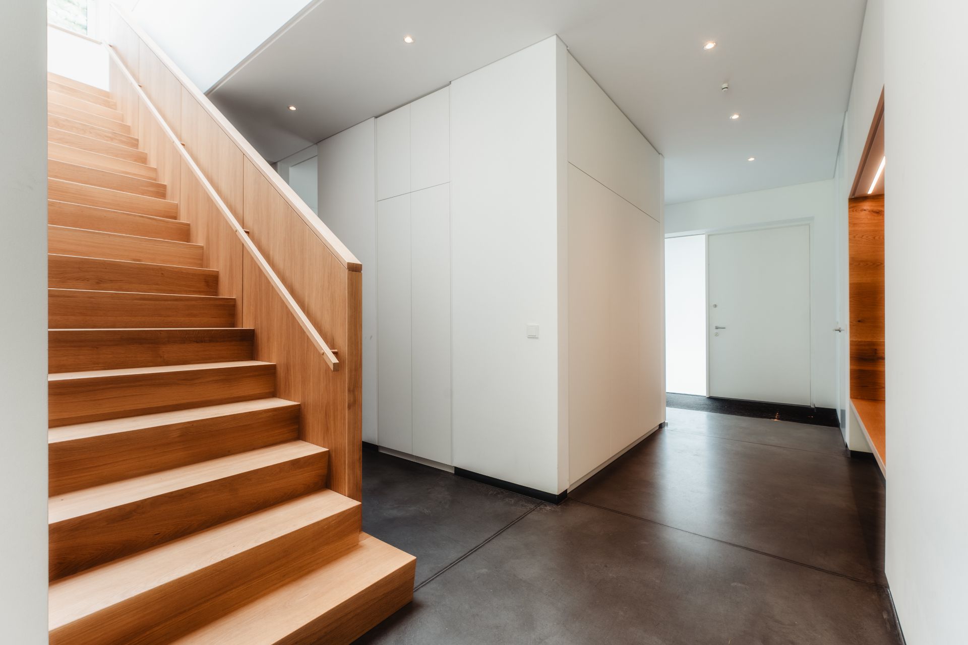 Heller Flur mit Holztreppe, weißen Wänden und dunklem Boden in einem modernen Haus.