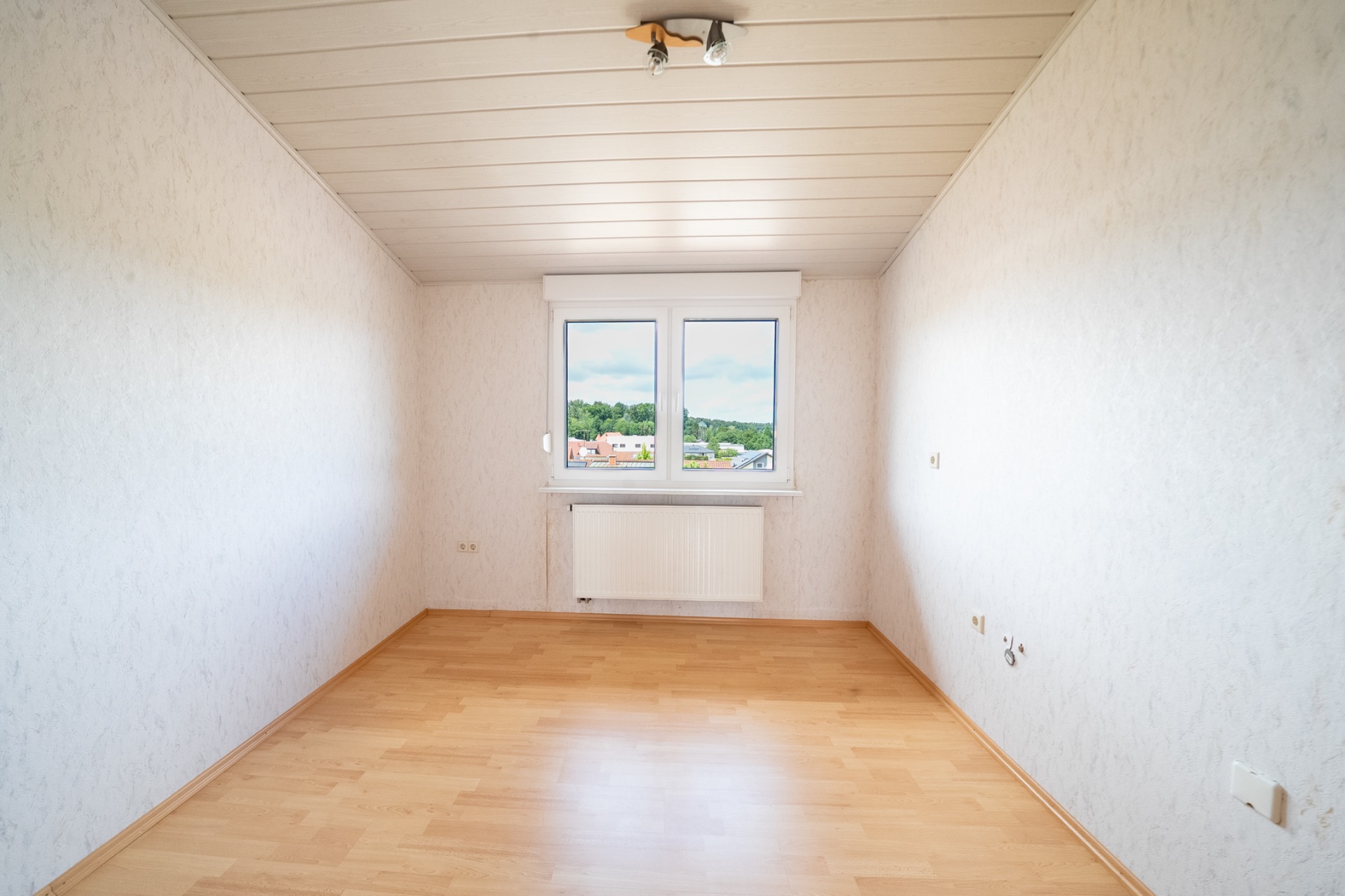 Leeres Zimmer mit Holzfußboden, zwei Fenstern und Heizkörper unter dem Fenster.