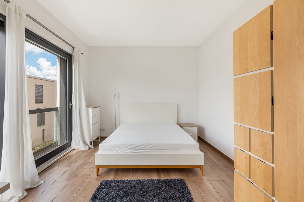Helles Schlafzimmer mit großem Fenster, Doppelbett, Schrank und Nachttisch auf Holzfußboden.