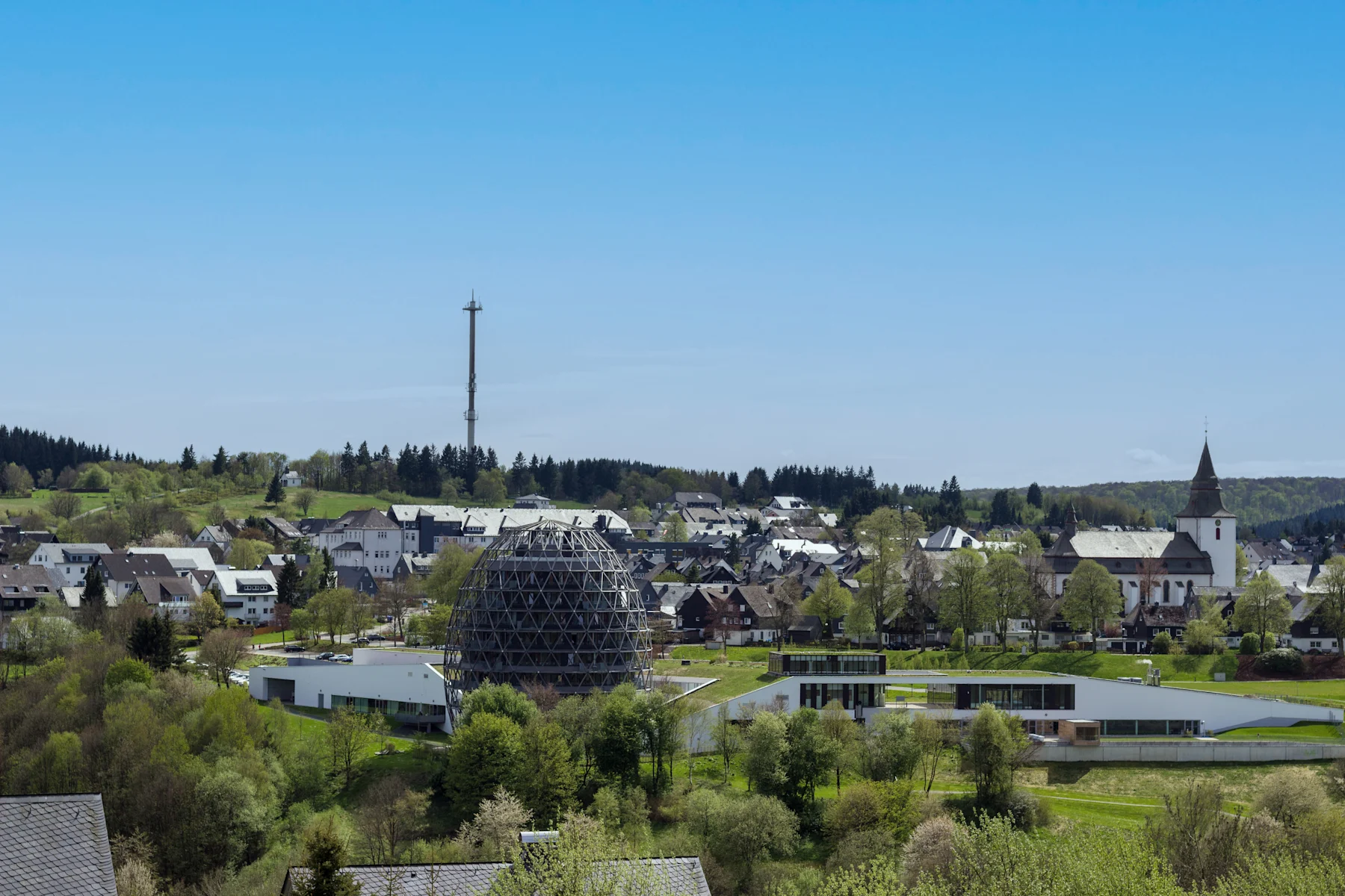 Weitläufige Landschaft mit Hügeln, Feldern, Wald und einem Dorf im Tal unter blauem Himmel.