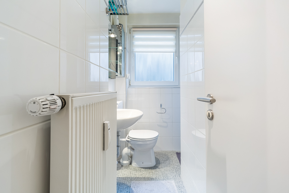 Helles Badezimmer mit Toilette, Waschbecken, Heizkörper und Fenster mit Sichtschutz.