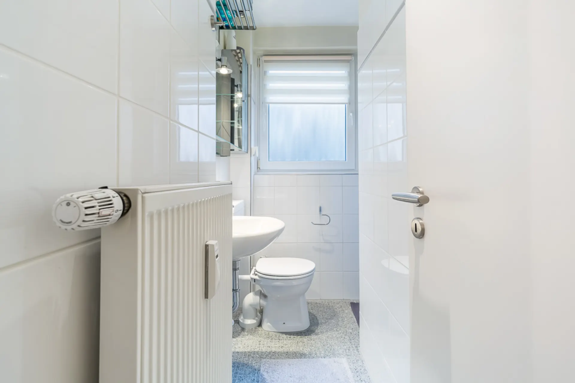 Helles Badezimmer mit Toilette, Waschbecken, Heizkörper und Fenster mit Sichtschutz.