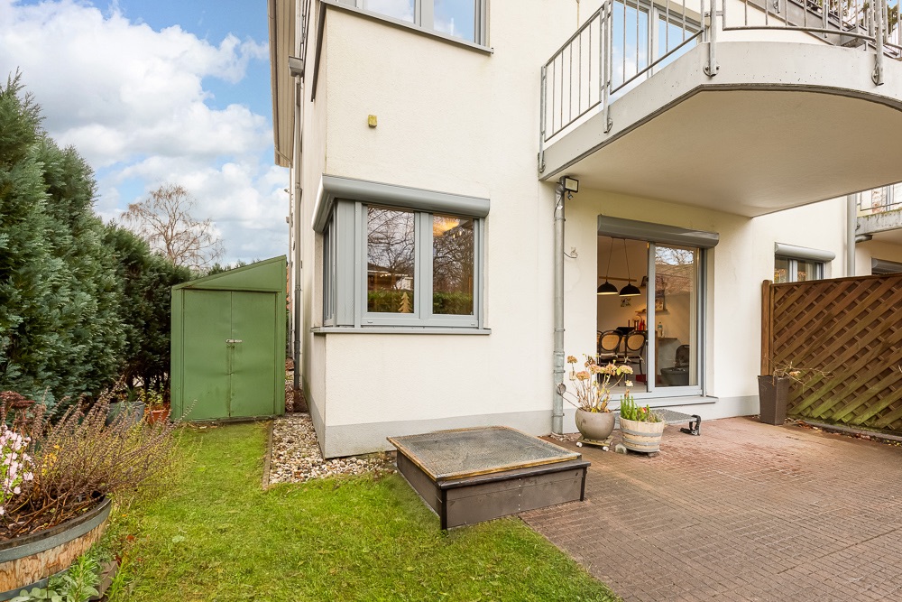 Erdgeschosswohnung mit Terrasse, Pflanzenkübeln und kleinem grünen Geräteschuppen im Garten.