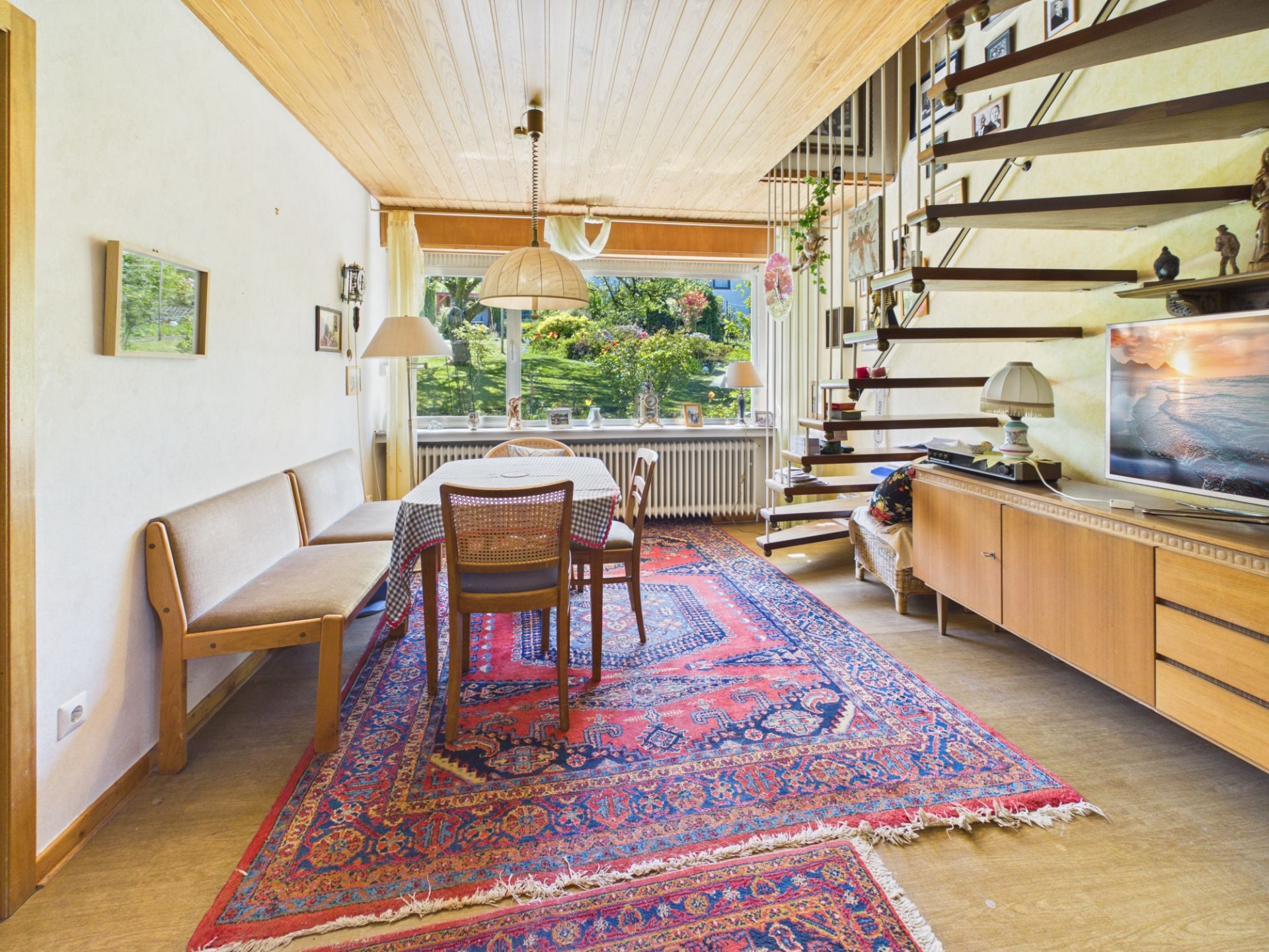 Wohnzimmer mit Teppich, Esstisch, Stühlen, Holzdecke, Sideboard, Fernseher und offener Treppe.