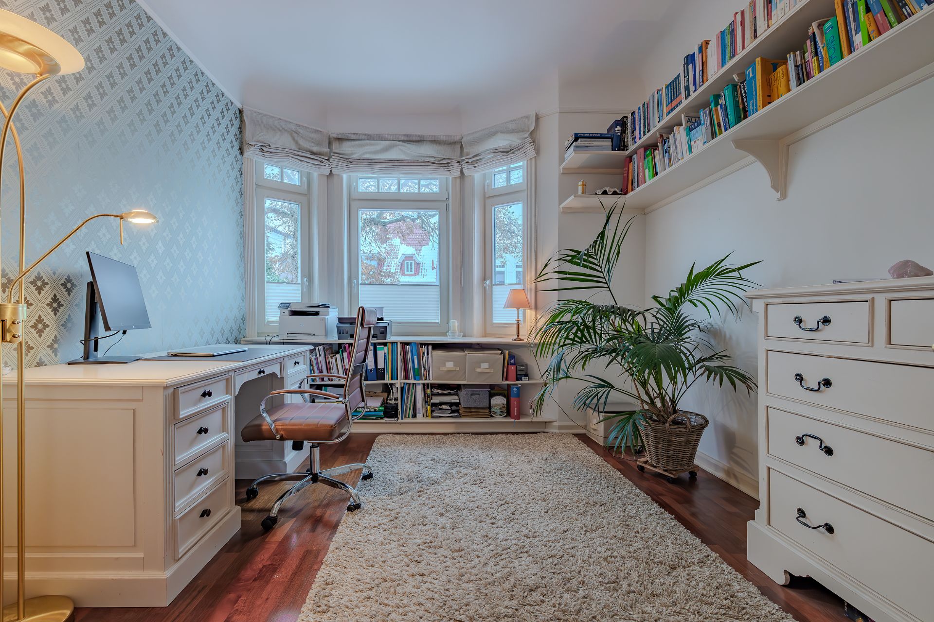 Helles Arbeitszimmer mit Schreibtisch, Bücherregalen, Teppich und großen Fenstern.
