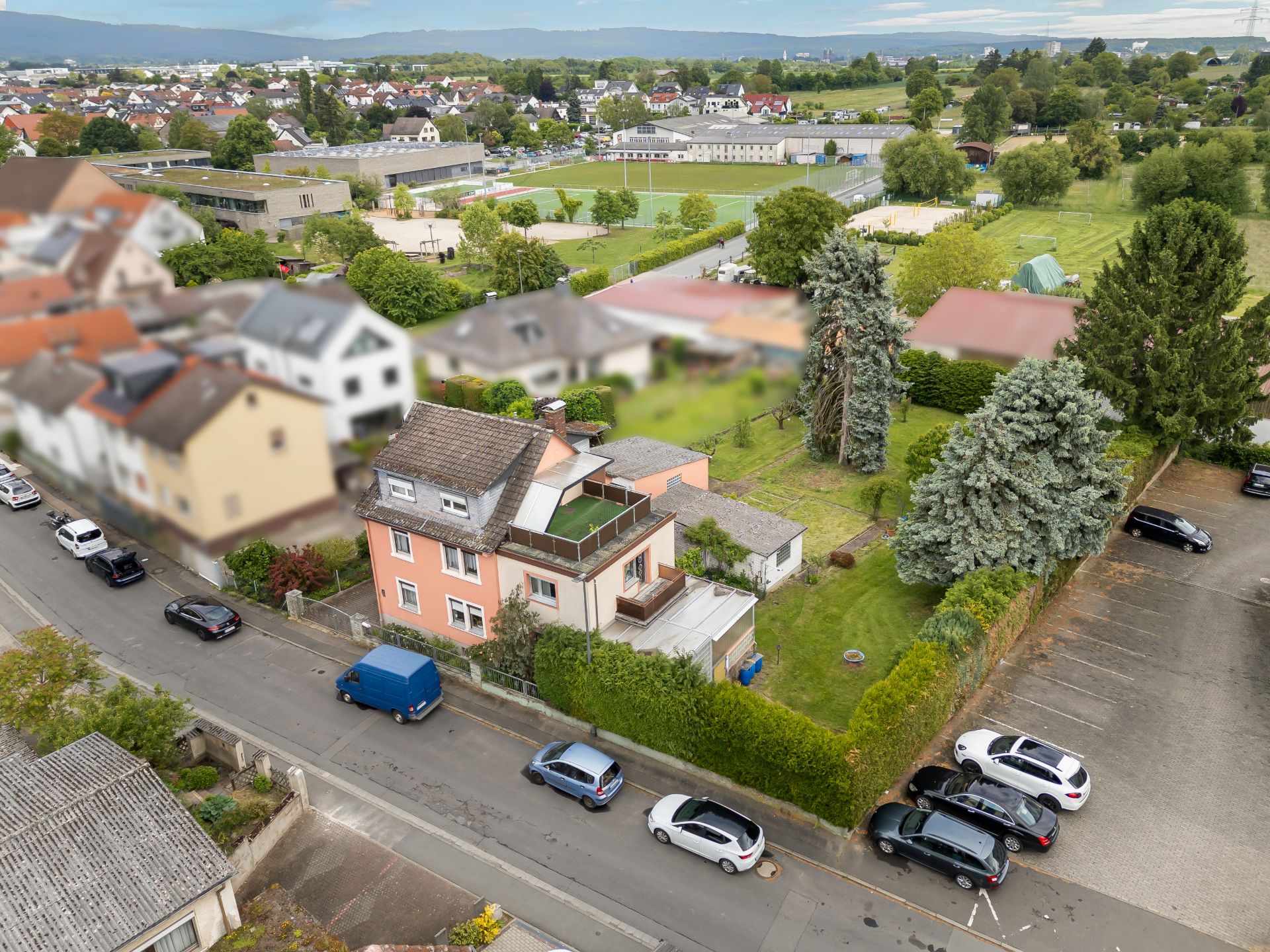 Luftaufnahme eines Wohnhauses mit Garten, angrenzendem Parkplatz und Bäumen in einer Wohngegend.