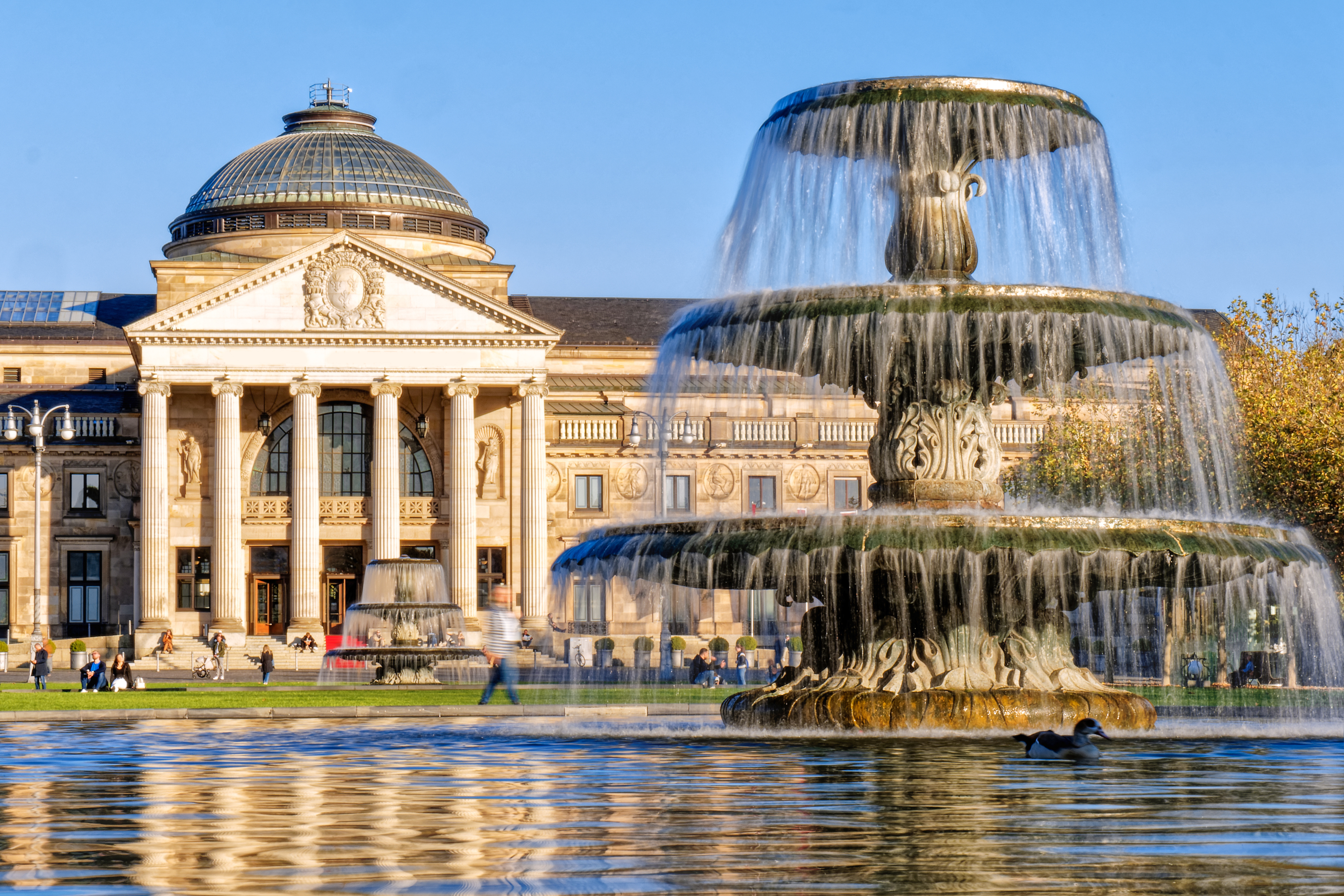 Großer Springbrunnen vor einem historischen Gebäude mit Säulen und Kuppeldach bei Tageslicht.