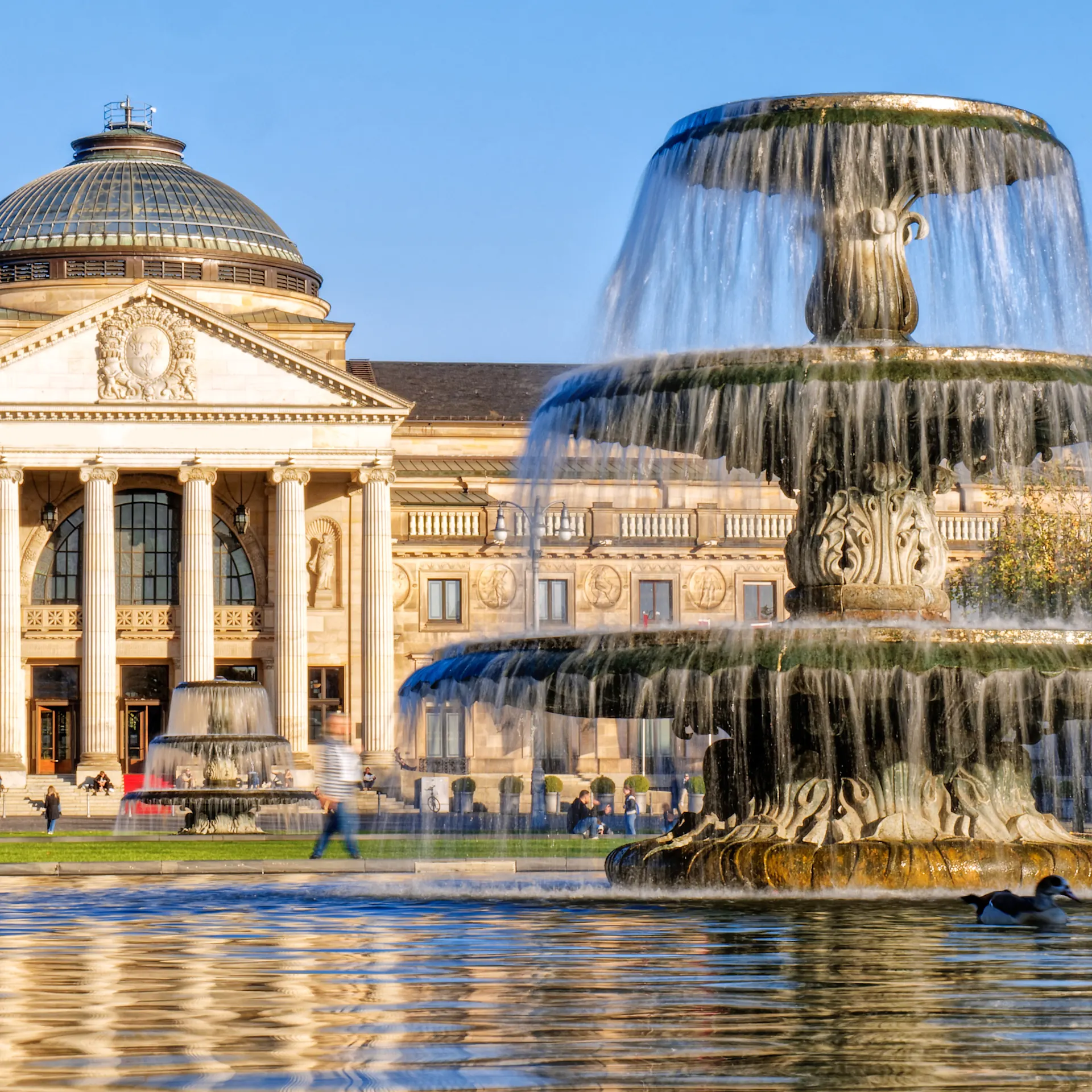 Großer Springbrunnen vor einem historischen Gebäude mit Säulen und Kuppeldach bei Tageslicht.