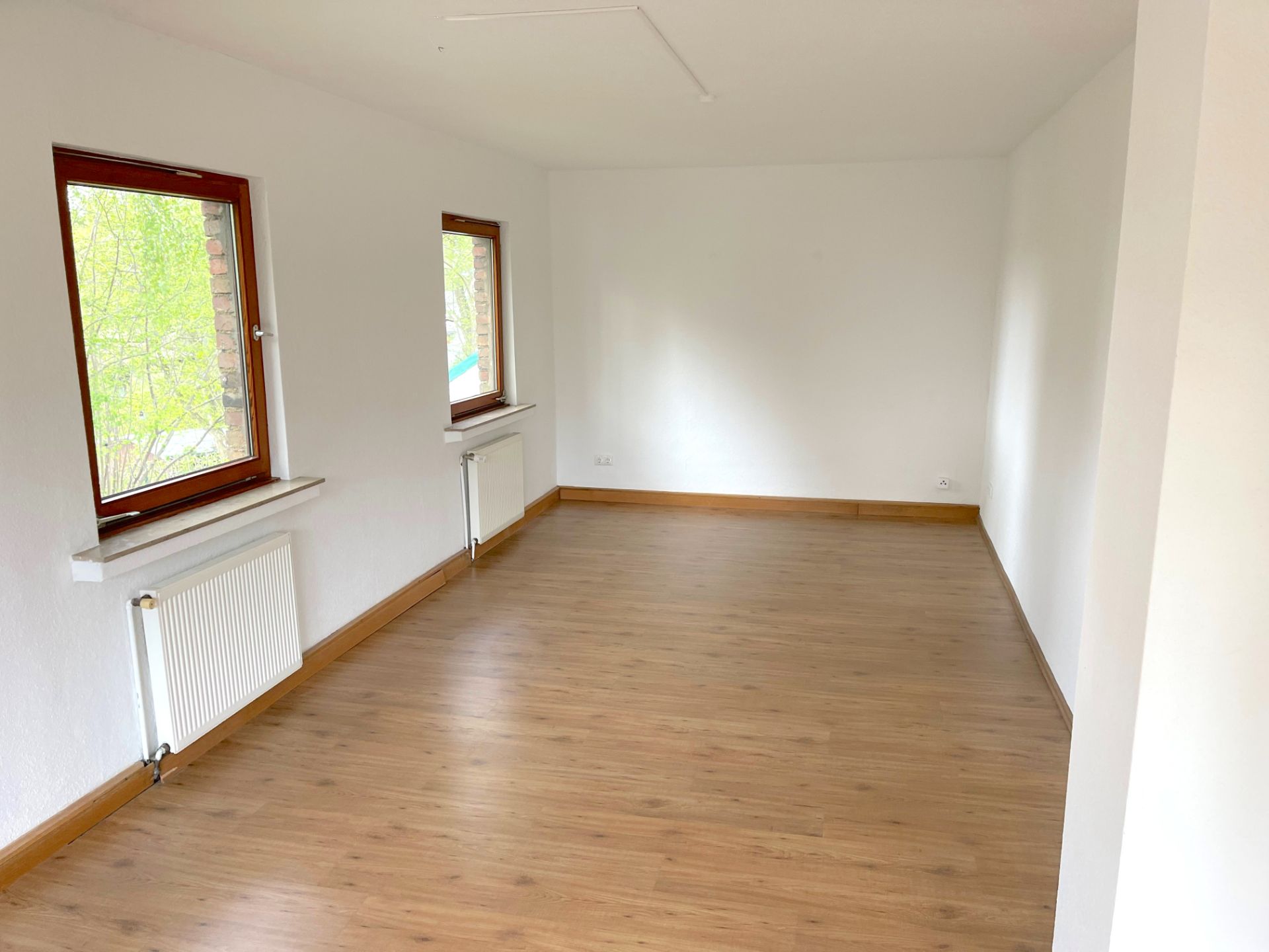Heller leerer Raum mit zwei Fenstern, weiß gestrichenen Wänden und Holzfußboden.
