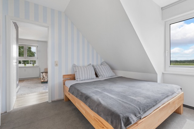 Schlafzimmer mit Holzbett, gestreifter Wand und Fenster mit Blick ins Grüne.