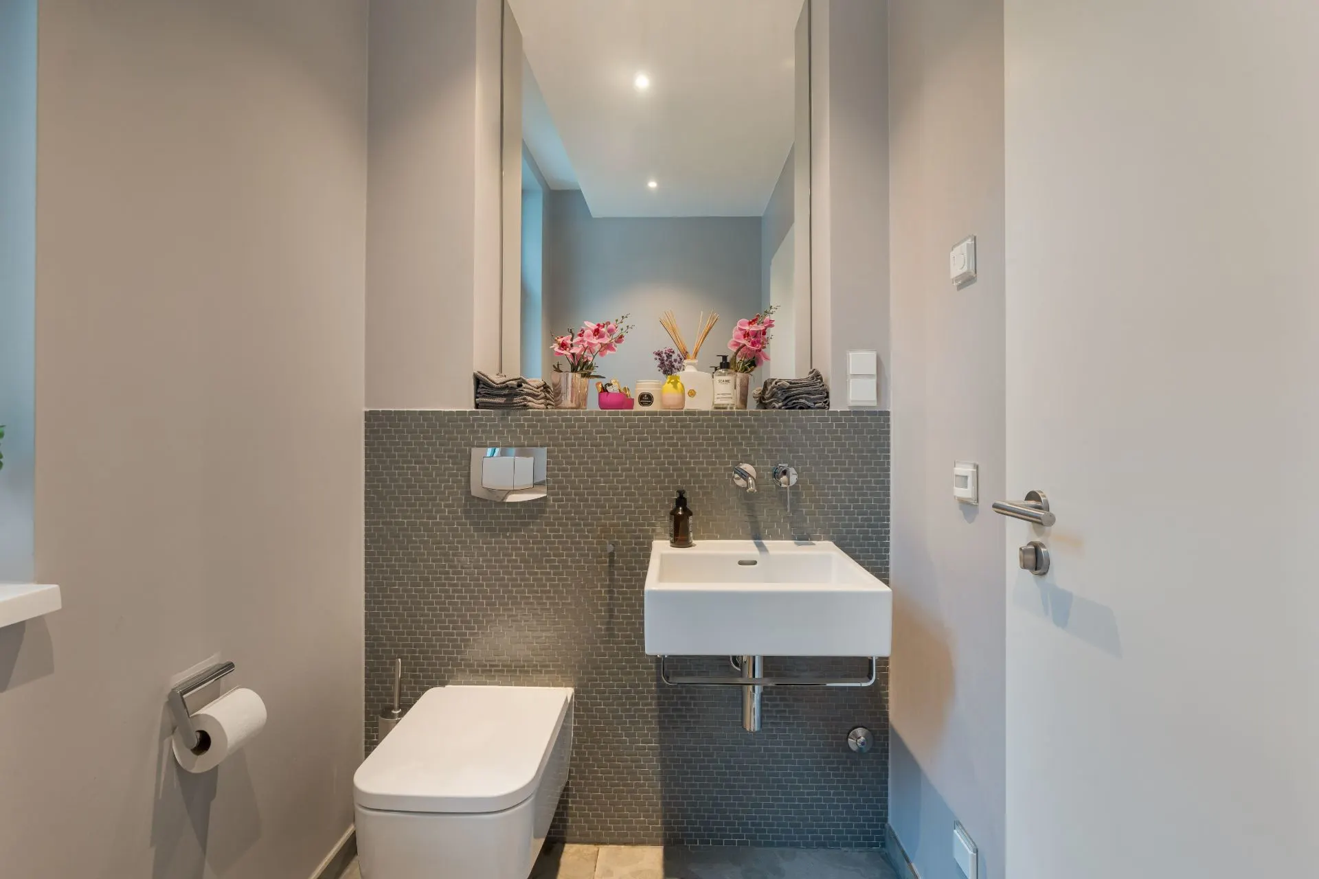 Modernes Badezimmer mit Wandtoilette, Waschbecken, Spiegel und Dekorationen auf einem Regal.