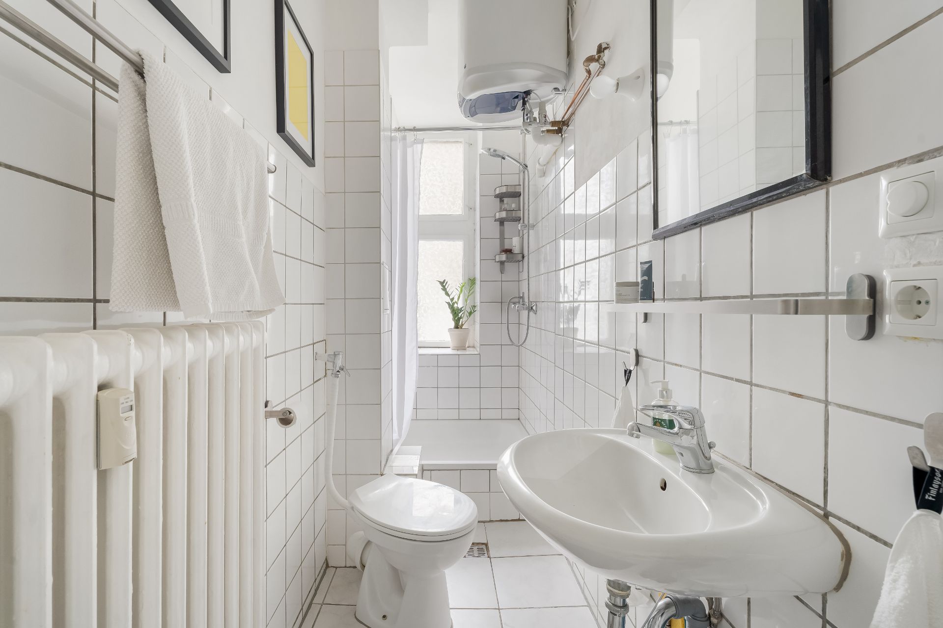 Helles Badezimmer mit Waschbecken, Toilette, Dusche und Fenster, weiße Fliesen an den Wänden.