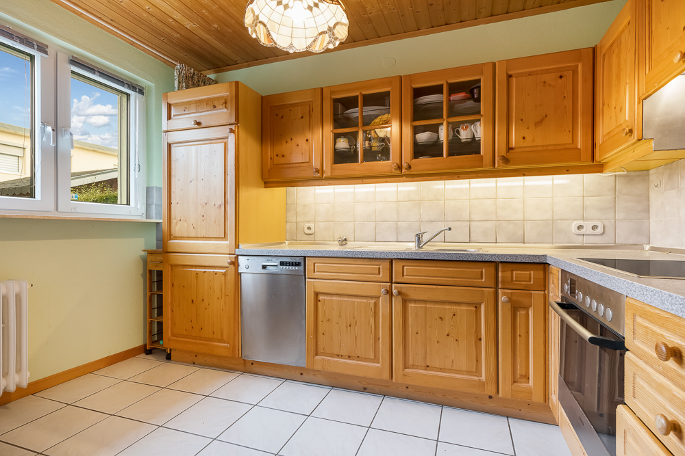 Helle Einbauküche mit Holzschränken, Spülmaschine, Fenster und Fliesenboden.