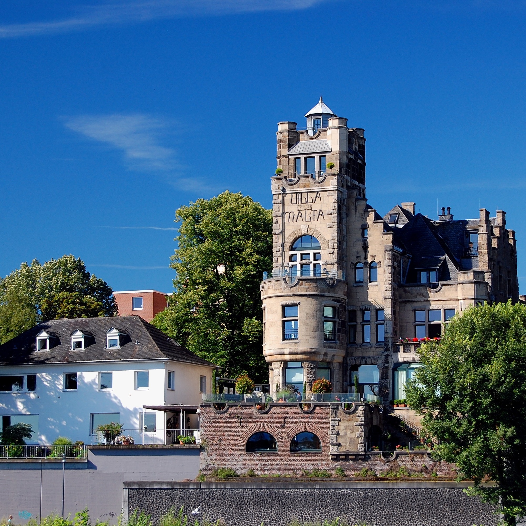 Großes historisches Wohnhaus mit Türmen neben einem weißen modernen Wohngebäude, Bäume im Vordergrund.
