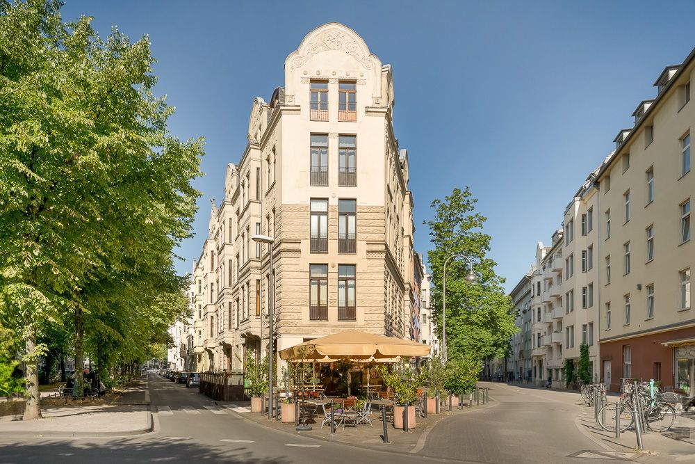 Eckhaus mit mehreren Etagen und Café mit Sonnenschirm im Erdgeschoss, umgeben von Straßen und Bäumen.