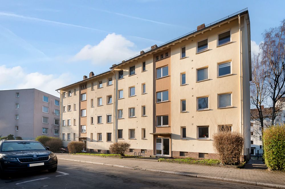 Mehrstöckiges Wohnhaus mit vielen Fenstern und einem Eingang, davor parkt ein Auto.