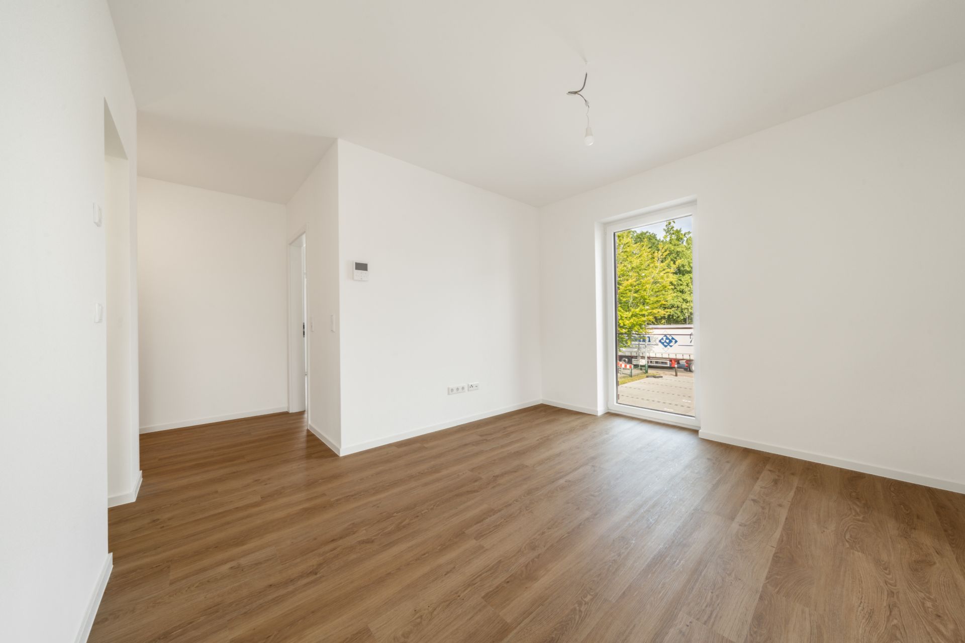 Leerer Raum mit Holzfußboden, weißen Wänden und Tür zu einem Balkon mit Blick ins Grüne.