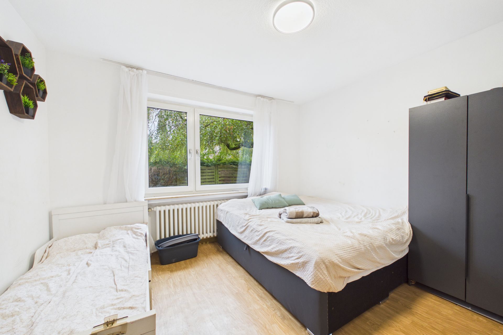 Helles Schlafzimmer mit zwei Betten, einem schwarzen Schrank und Fenster mit weißen Vorhängen.