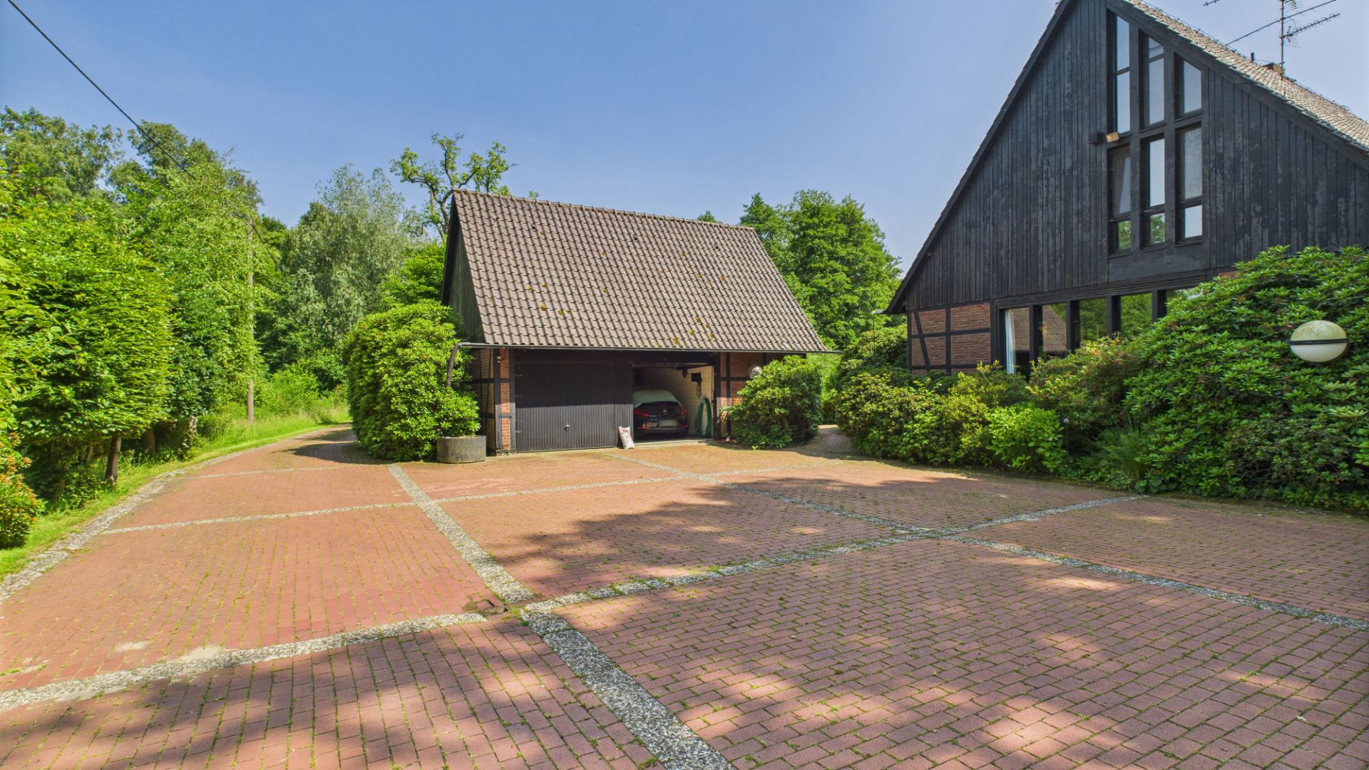 Gepflasterter Hof mit Carport und zwei Wohngebäuden, umgeben von Grün und Bäumen.