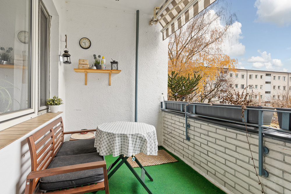 Balkon mit Holzbänkchen, rundem Tisch mit Tischdecke, Regal und Pflanzen, Blick auf Wohngebäude.