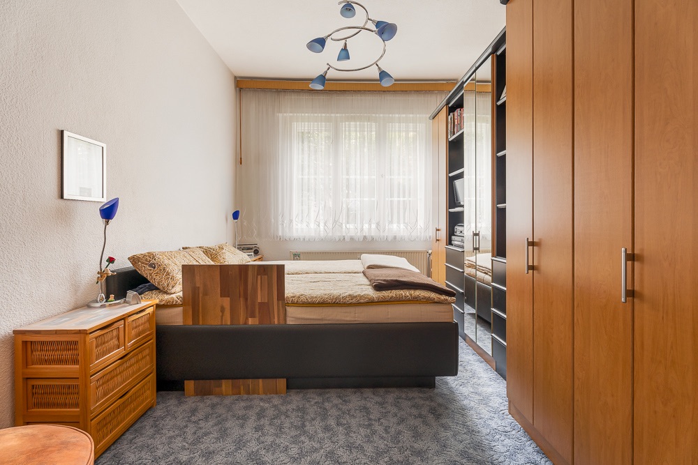 Schlafzimmer mit Doppelbett, Holzmöbeln, Teppichboden und großem Fenster mit Vorhängen.