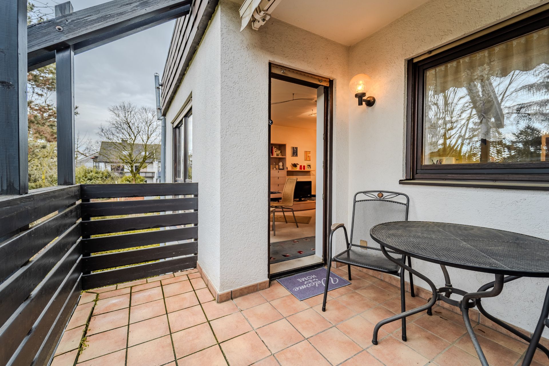 Kleine Terrasse mit Terrassentisch und zwei Stühlen, Tür zum Wohnzimmer, Fenster mit Gardine.